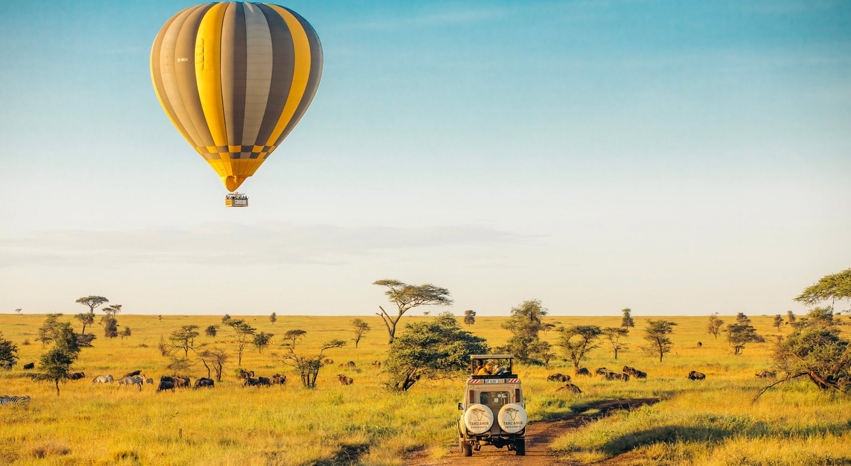 Ein Heißluftballon schwebt über der Serengeti, während darunter ein Fahrzeug von Tansania-Experience durch die weite Landschaft fährt.