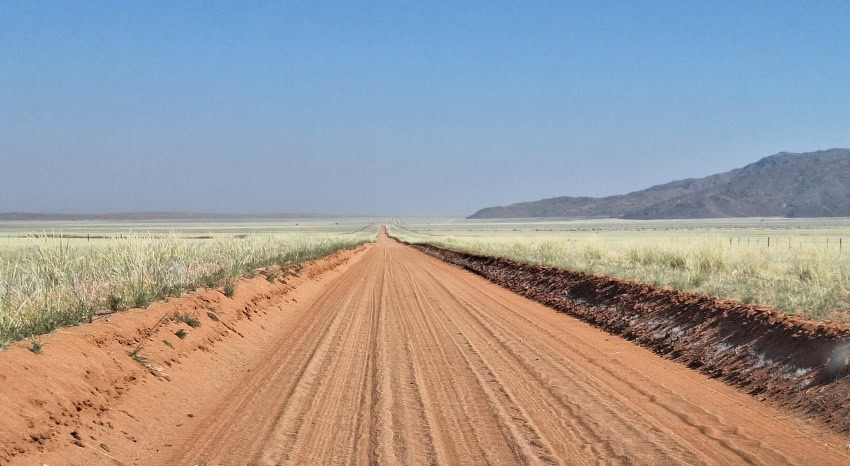 Sandpiste durch die Savanne Namibias mit bergiger Landschaft auf der rechten Seite.